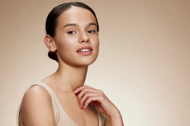 Young woman with natural makeup, wearing neutral clothing, exuding confidence and tranquility against a soft beige background
