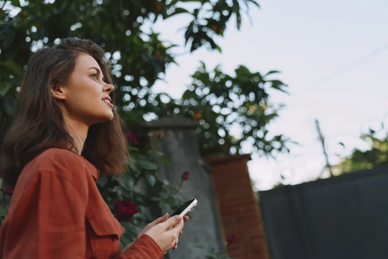 Woman lifestyle portrait outdoors, relaxed casual pose, holding smartphone, looking up with thoughtful smile, natural daylight, everyday scene, candid moment
