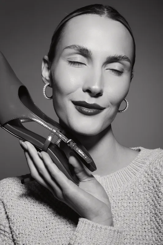 Caucasian woman with fair skin and closed eyes holds red high heel shoe near her face, wearing knitted sweater and hoop earrings in black and white studio portrait.