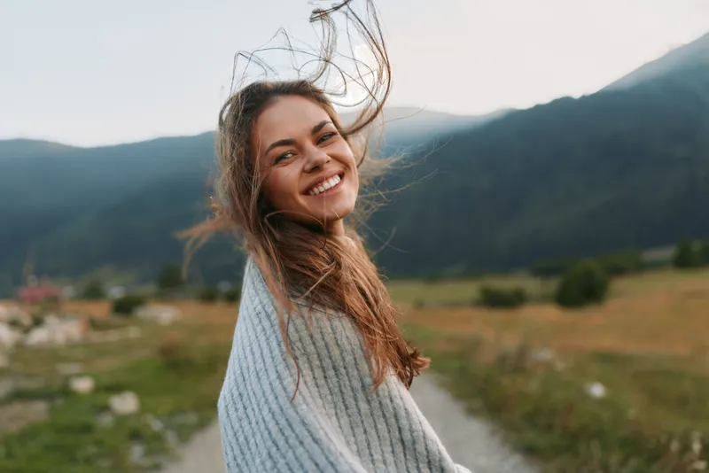 Happy woman outdoors smiling with hair blowing in wind wrapped in cozy sweater natural light scenic mountain landscape background