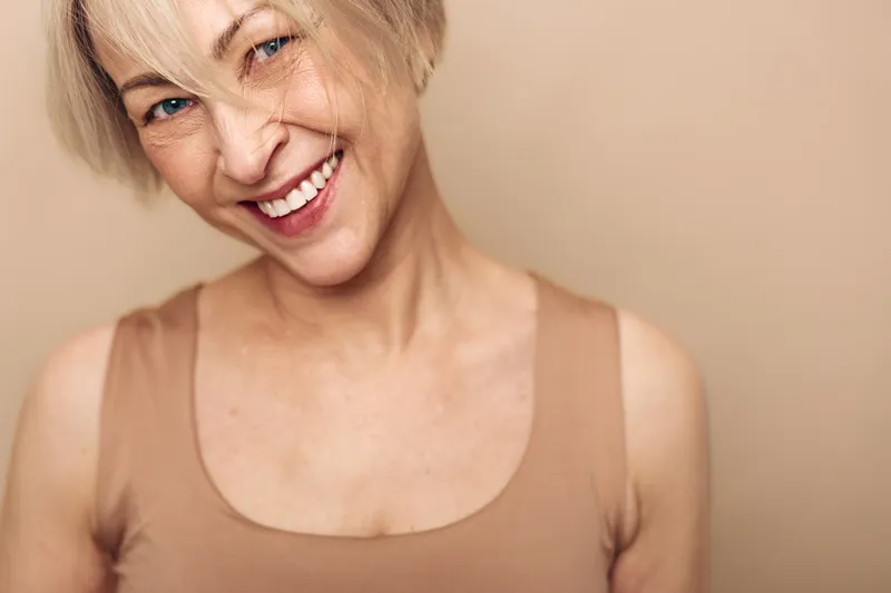 Smiling mature woman with short blonde hair wearing beige tank top looks joyful against a neutral background. Concept of happiness, confidence, aging gracefully and positive emotions.