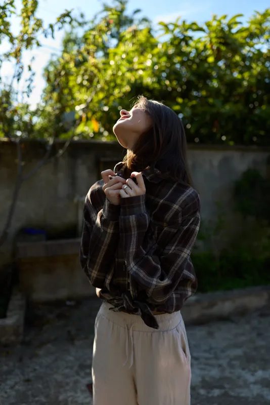 Young woman outdoors wearing a plaid shirt poses with lifted face toward the sun, showcasing casual lifestyle and natural light, relaxed mood in a sunny garden setting