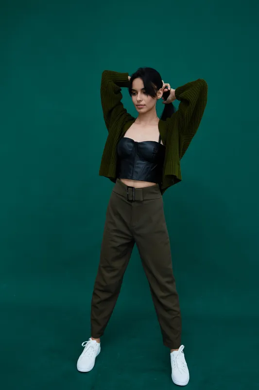 Young woman posing confidently in black leather top and olive pants against green background, wearing knitted cardigan and white sneakers in casual style.