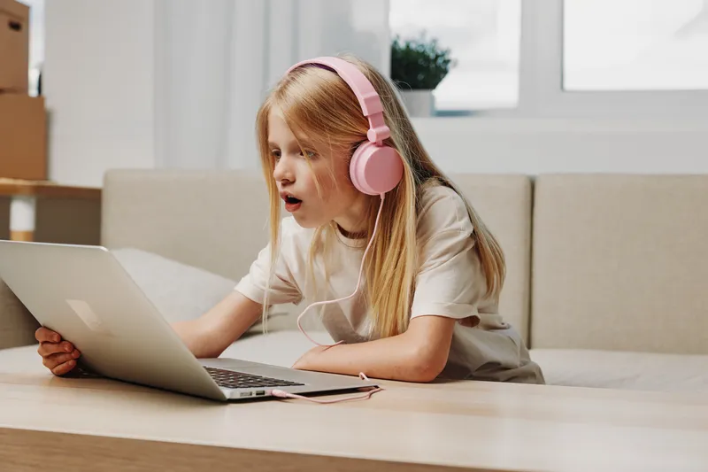 Curious blonde girl with pink headphones uses laptop at table in bright modern room, engaged in digital learning or entertainment.