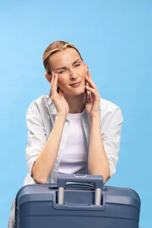 Stressed woman talking on phone holds head with fingers, sits behind suitcase on blue background. Concept of travel anxiety, frustration, and emotional discomfort during trip.