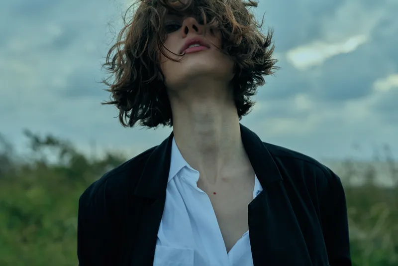 Young woman with tousled hair, blazer and white shirt, looking up under a cloudy sky in an open outdoor setting, moody fashion shot