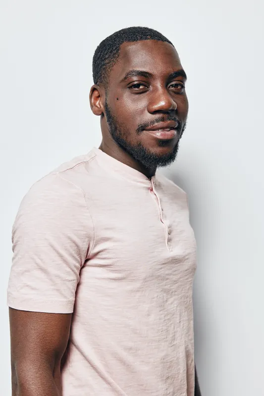 African American man with dark skin and short hair wearing light pink shirt standing against white background looking confident and relaxed in studio portrait.