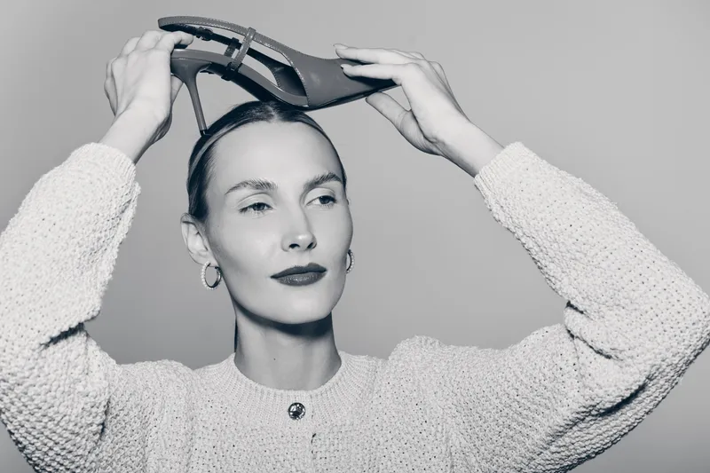 Young white woman with pale skin puts high heel shoe on head wearing textured sweater in studio. Concept for fashion and style.