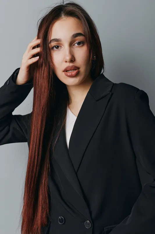 Young woman with long brown hair wearing black blazer and white shirt posing against gray background. Portrait of confident female with natural makeup and relaxed expression indoors.