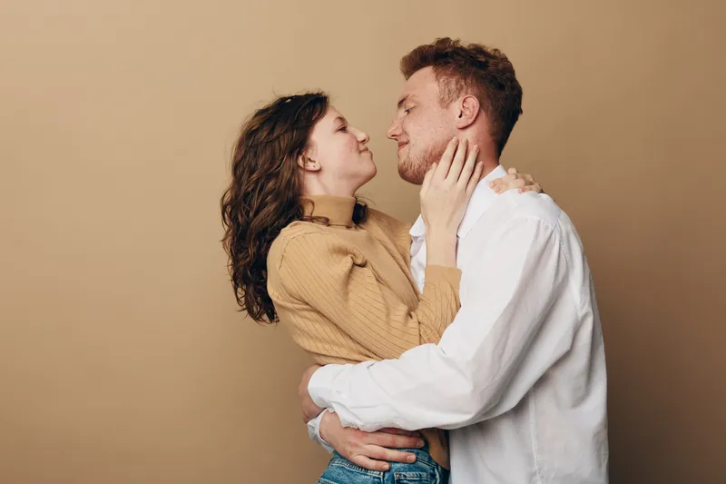 Young couple embraces and looks into each other's eyes with affection. Woman in beige sweater and man in white shirt share intimate moment on neutral background. Concept of love and connection.