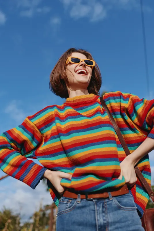 Young woman in a rainbow sweater wearing yellow sunglasses, smiling with hands on hips in a casual outfit under blue sky outdoors, joyful travel and lifestyle portrait.