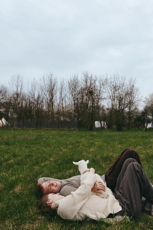 Casual outdoor lifestyle scene with a couple lying on grass, cuddling a young goat kid under a gray sky, conveying warmth, tenderness, and simple joy in nature.