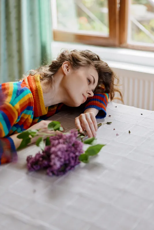A woman rests her head on a table near a vase of lilac flowers, wearing a colorful striped sweater. A quiet intimate moment capturing fatigue, solitude, and home life.