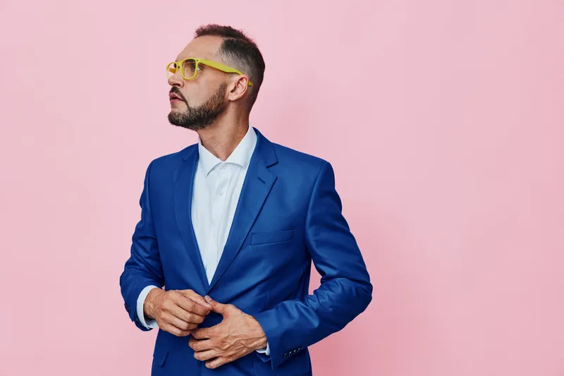 A professional man in a blue suit stands against a soft pink background, projecting confident focus, calm demeanor, and poised posture for a modern business portrait.