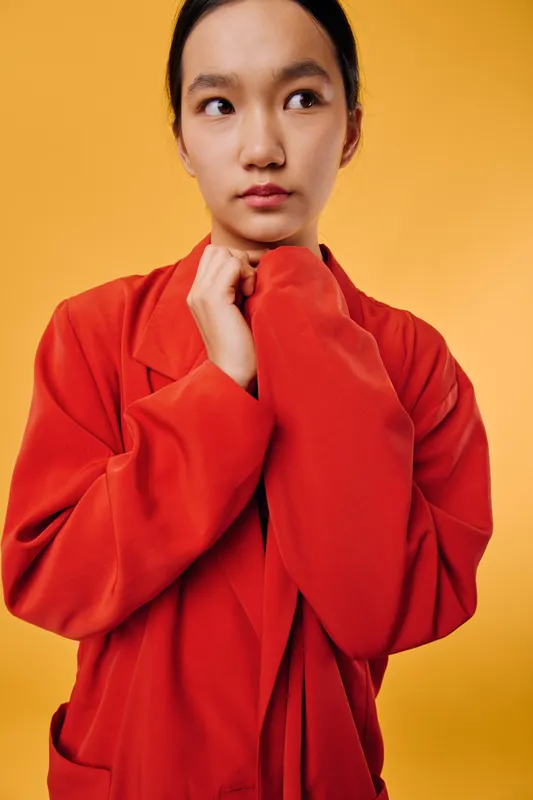Young woman with contemplative expression wearing oversized red coat standing against orange background with hands clasped near face, casual portrait with soft lighting.