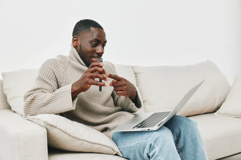 Young Black man engaging in a video call at home, wearing a cozy sweater, sitting on a sofa, smiling with an open laptop, showcasing remote communication