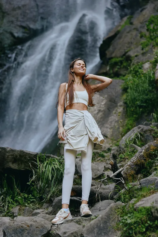 woman standing near waterfall, dressed in white activewear, enjoying nature outdoors with a serene expression, surrounded by rocks and greenery, sunny weather