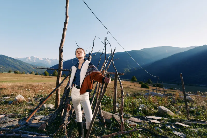Woman in jacket and boots standing by a rustic fence in a mountain field, fashion portrait with sunlight and smile, countryside landscape, serene valley view and outdoor lifestyle scene.