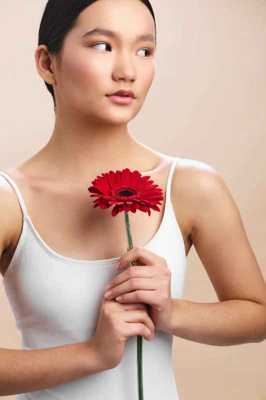 asian woman holds vibrant red gerbera flower in a simple studio setting, calm gaze to the side, soft lighting and clean background create an elegant, serene portrait.