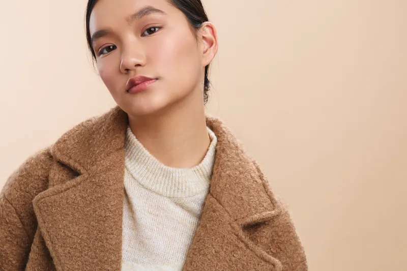 asian woman with a calm expression wears a soft beige coat and knit sweater, posed against a neutral backdrop, conveying warm elegance and thoughtful presence.