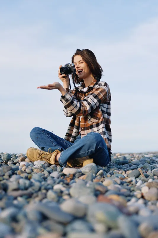 Woman photographer sits on a pebble beach in casual plaid shirt and jeans, capturing moments with a camera under bright skies, showcasing outdoor lifestyle, creativity, focus, and connection with