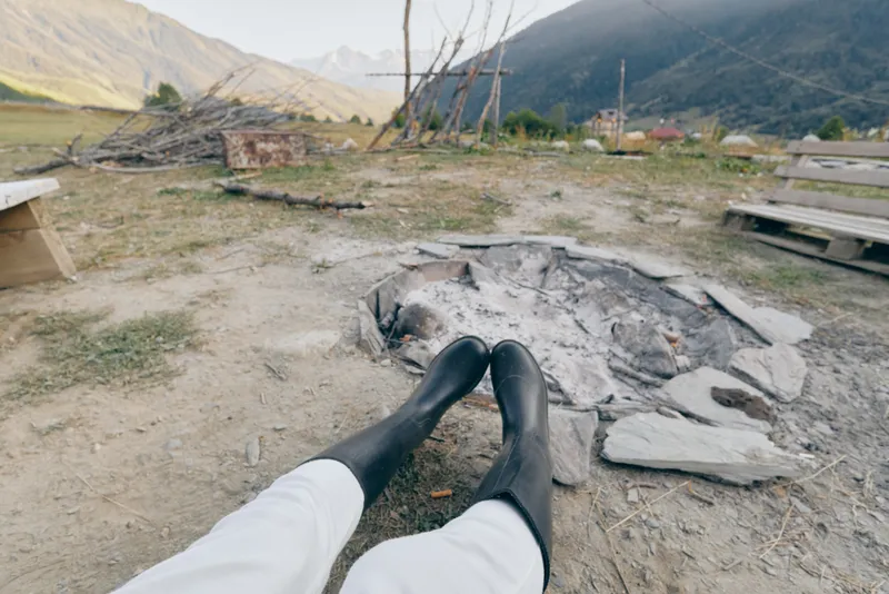 Boots resting near a cold campfire pit with distant mountains in the background, outdoor legs stretched out on rough countryside ground, quiet relaxation after a long hiking day.