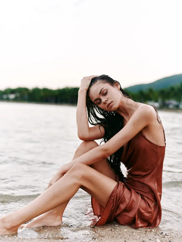 Young woman sits on a sandy beach by calm water, wearing a rust brown dress. Wet hair frames a thoughtful face, creating a serene, intimate moment in natural light.