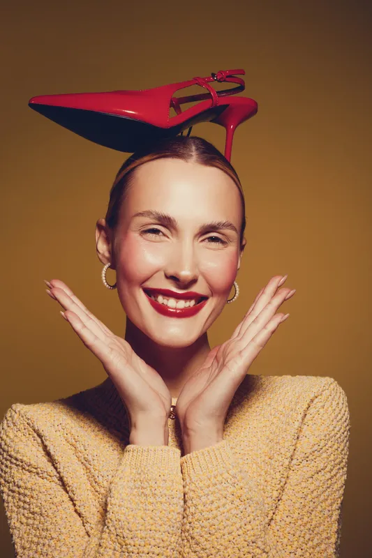 Smiling woman with red high heel shoe balanced on her head, wearing yellow sweater and gold hoop earrings. Concept of fashion, fun, and playful portrait with warm lighting.
