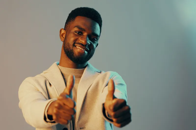 Cheerful African man giving thumbs up with a smile, wearing light colored casual clothing, showcasing positivity and confidence in a modern studio setting