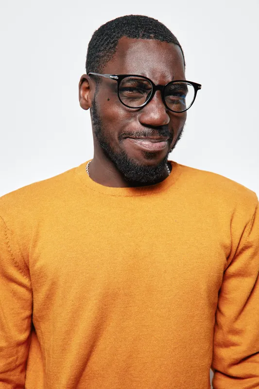 African man with dark skin wearing glasses and orange sweater smiling with closed eyes on white background. Portrait of happy black male with beard and short hair in casual clothing.