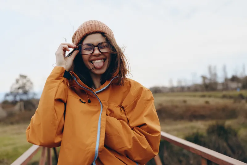 Woman lifestyle outdoors, smiling in orange raincoat and beige beanie, glasses visible, talking on phone for communication, laughter and relaxed stance on a rural bridge with countryside field