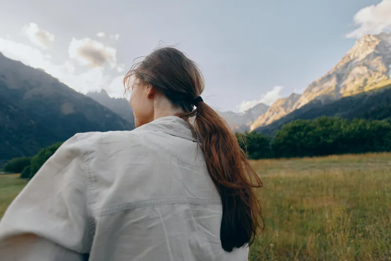 A woman with a light shawl stands with her back to the camera, gazing at rugged mountains and a green valley, capturing a calm, exploratory outdoor moment.