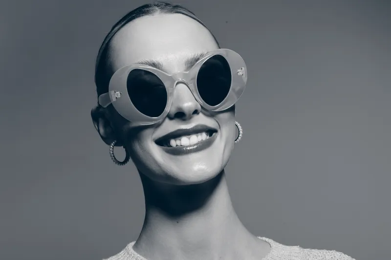 Smiling white woman with fair skin wearing round sunglasses and hoop earrings on gray background.