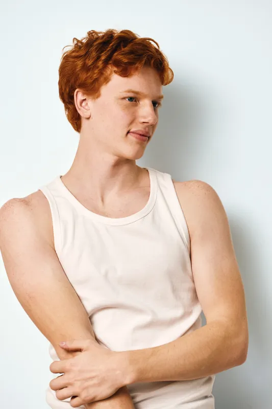 Young man with red hair and fair skin wearing white tank top posing with folded arms, looking away on plain white background in casual style for portrait or fashion.