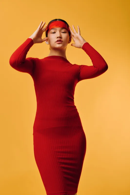 Young woman in red ribbed dress with red headband posing with eyes closed and hands near face on orange background. Fashion, portrait, style, and minimalistic studio shot.