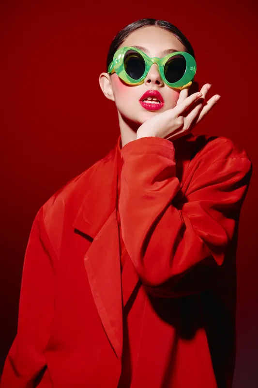 Young woman in oversized green sunglasses and bright oversized red coat poses against crimson background. Stylish fashion portrait expressing boldness and creative beauty concept.