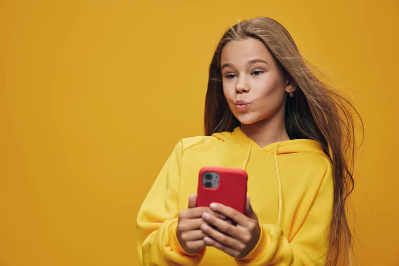 Teenage girl in yellow hoodie using red smartphone with focused expression and long hair against orange background, technology and youth culture concept with casual style.