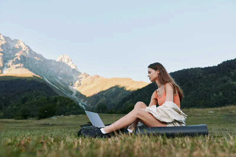 A relaxed young woman sits on a yoga mat with a laptop in a scenic meadow, enjoying sunny weather and a mountainous backdrop, creating a calm, productive outdoor work atmosphere.