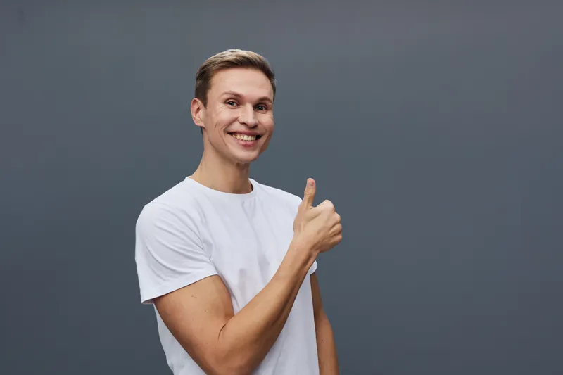 young smiling man thumb up positive emotion white t-shirt casual wear dark plain background studio portrait digital detox concept