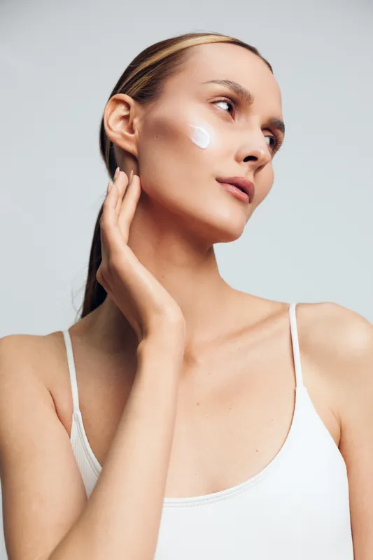 Young woman with clear skin applying facial cream on cheek, natural skincare routine, soft lighting, neutral background, focused expression, beauty and wellness concept