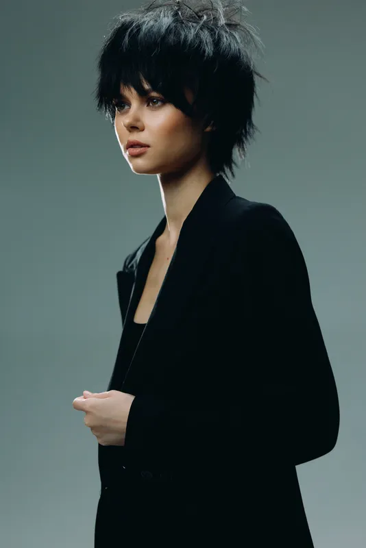 Young woman with short curly hair wearing black jacket posing in studio with neutral gray background.