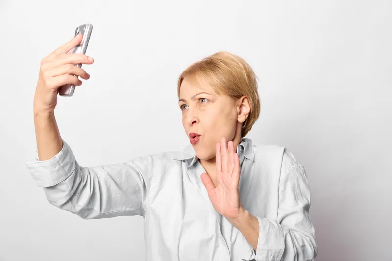 Surprised woman holding smartphone for a selfie, hand on cheek, shocked expression, indoor white background, conveying anxiety, fear, stress, communication.