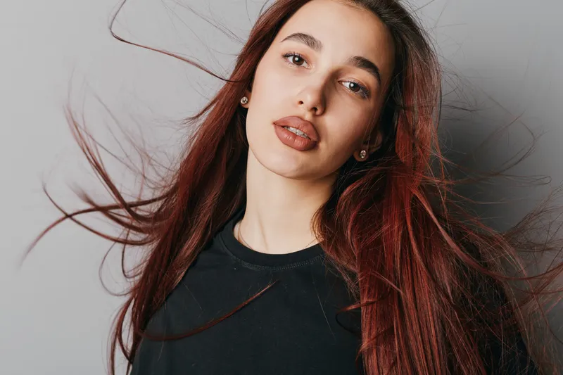 Young woman with long red hair and natural makeup posing in black shirt against gray background. Beauty portrait with flowing hair and calm expression in studio lighting.