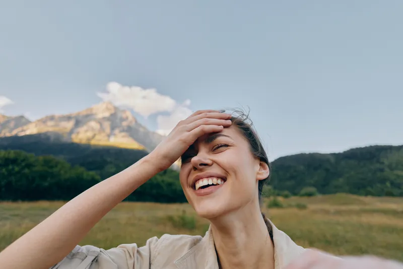 Young woman with brunette hair laughs joyfully while taking a selfie outdoors in a grassy field with mountains in the background under a clear sky.