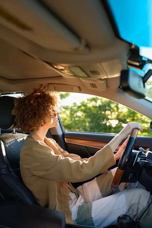 Woman driver sits in a vintage car, hands on the wheel, focused expression, casual outfit, retro film color palette, natural light through window, calm confident mood driving along a tree lined street