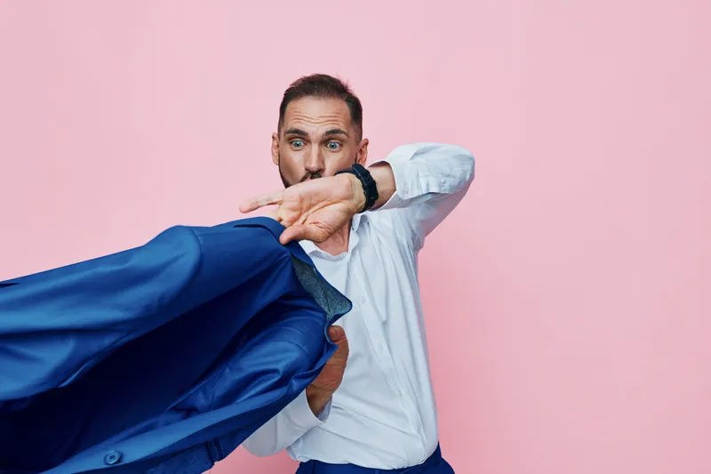 Professional man in a white shirt pulls a blue jacket across a bold pink background, conveying confidence, determination, and a moment of surprise in a corporate scene.