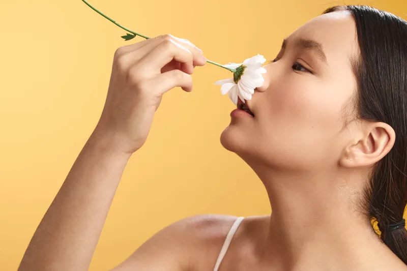 asian woman smelling a delicate flower in a bright studio environment, capturing a serene moment of calm appreciation, with soft warm light, gentle shadows, and a vibrant yellow background.