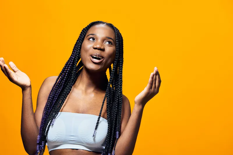 A joyful young Black woman with braided hair poses against a vibrant yellow background, expressing excitement and energy in a trendy outfit