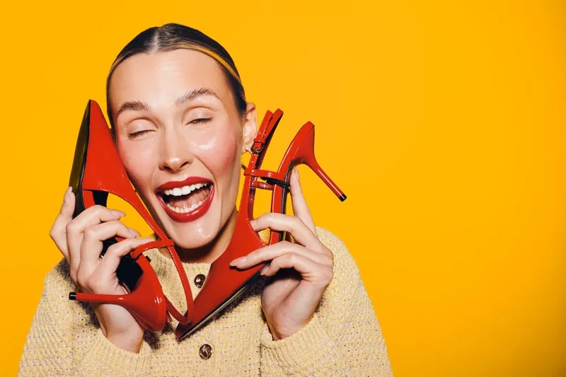 Happy woman with red lipstick holds red high heel shoes near face, smiling with closed eyes on bright yellow background. Concept of fashion, style, and joyful emotions.