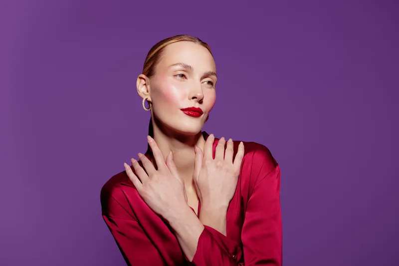Elegant woman with red lipstick and red blouse poses with crossed hands on purple background. Studio portrait with soft lighting highlights smooth skin and confident expression.